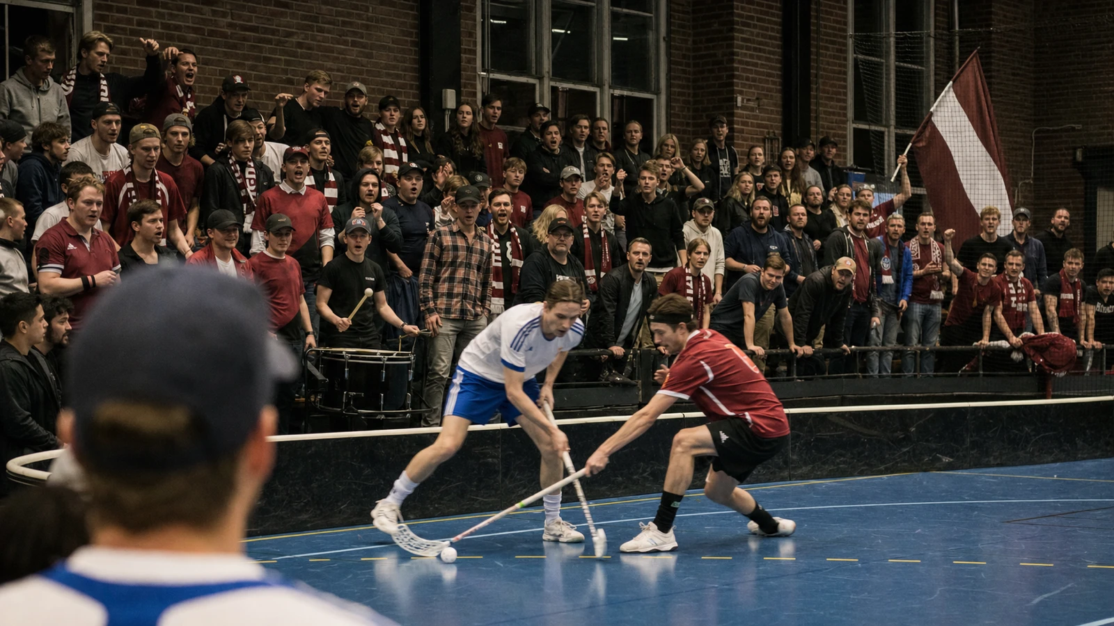 Supporterläktaren står upp med halsdukar och trumma medan två spelare kämpar om bollen närmast sargen